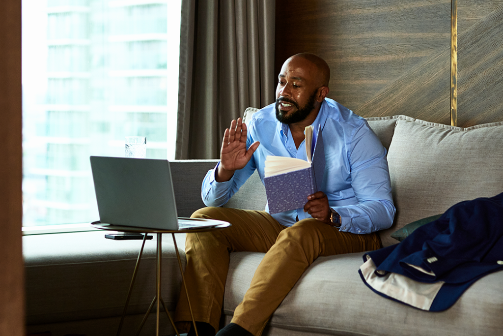 Homem negro sentado no sofá com um livro na mão e de frente para um notebook em cima de uma mesinha