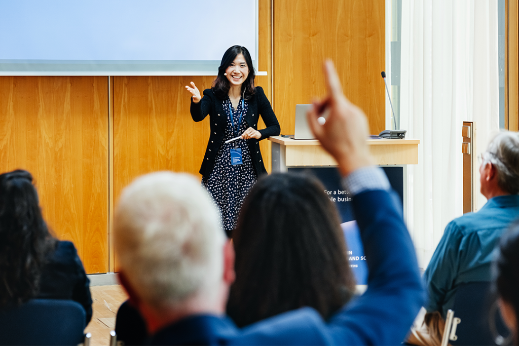 Mulher apresentando em uma sala de conferência, apontando e sorrindo para a audiência que levanta as mãos em participação.