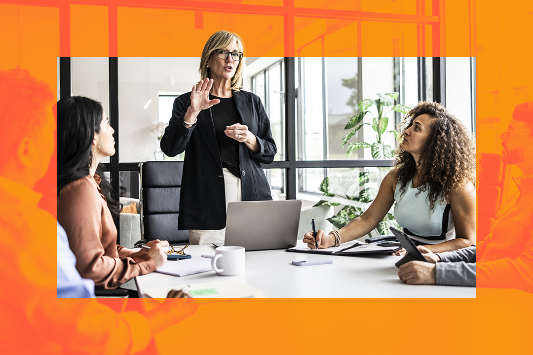 Executivos de uma empresa durante uma reunião. Uma moça loira está em pé e duas mulheres sentadas em uma mesa.