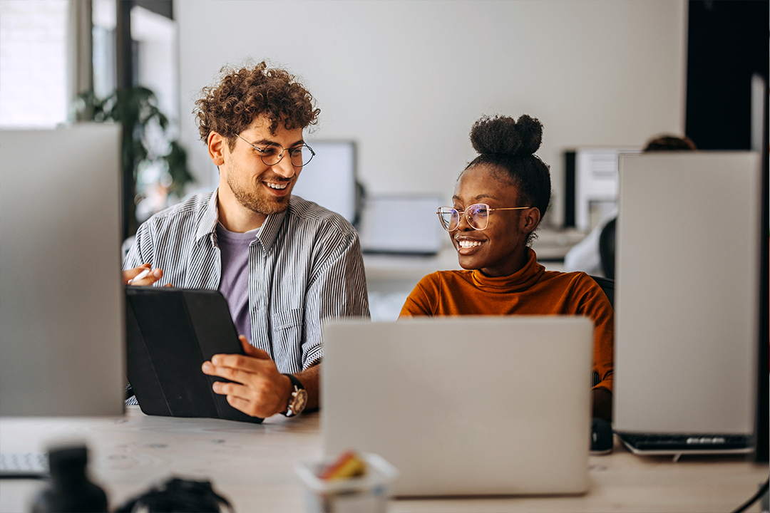 Homem e mulher sentados a uma mesa usando computadores e sorrindo