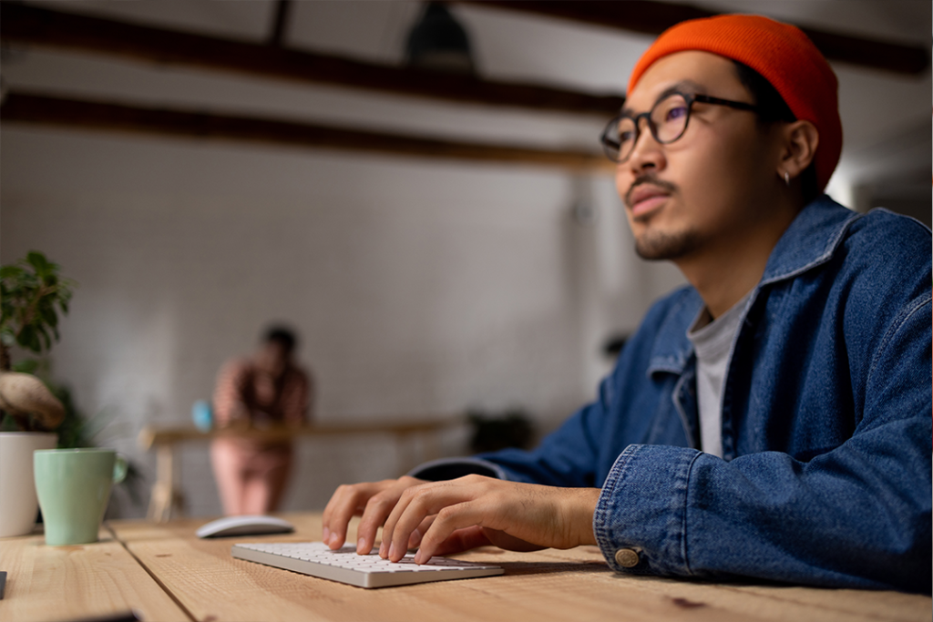 Homem com jaqueta jeans e gorro vermelho olhando com atenção para um notebook