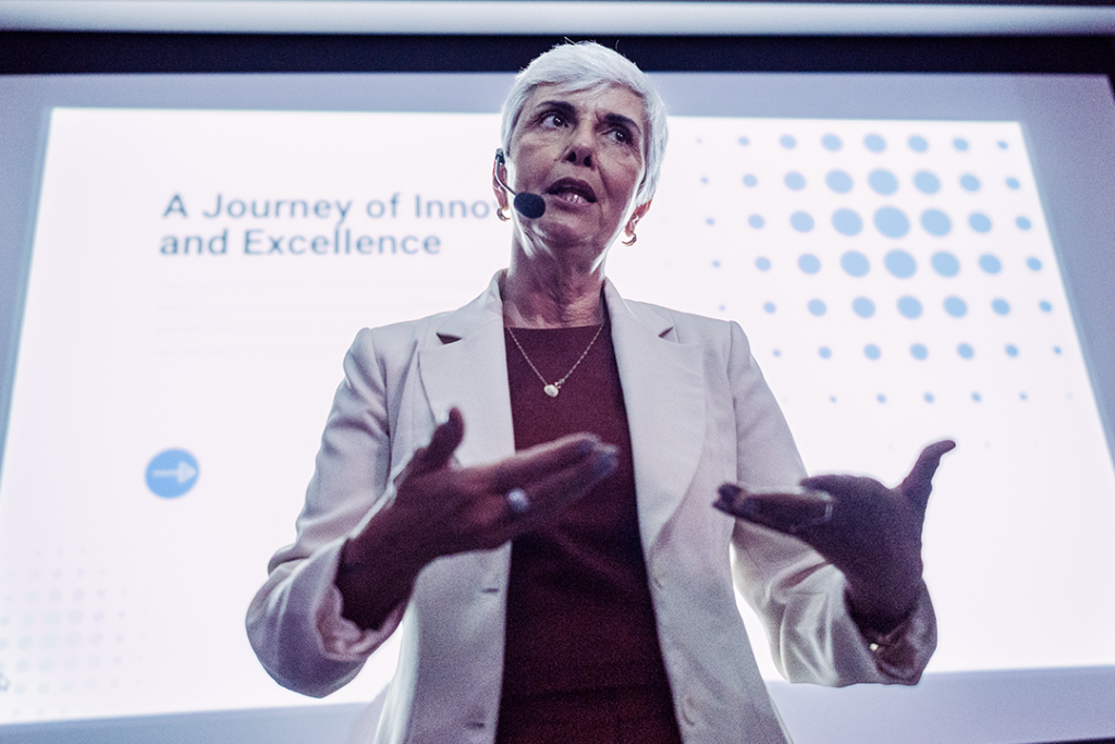 Mulher em palestra destacando a inovação e a excelência, com apresentação ao fundo sobre jornada de inovação. Ambiente profissional e inspirador.