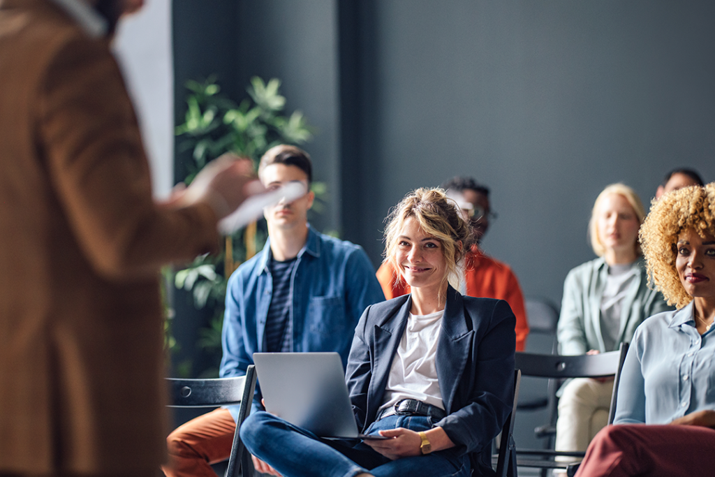 Imagem de mulheres e homens em um treinamento corporativo. Jovem mulher sorrindo com laptop em mãos, escutando o treinamento.