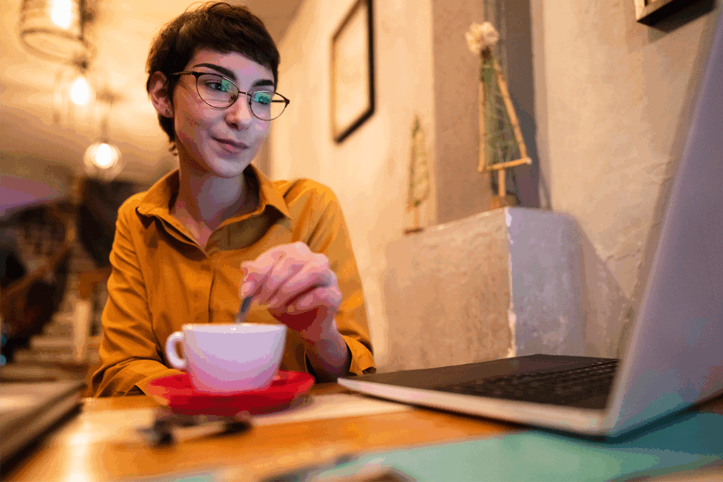 Mulher em frente a um computador e mexendo uma colher dentro de uma xícara de café, organizando as informações de um slide
