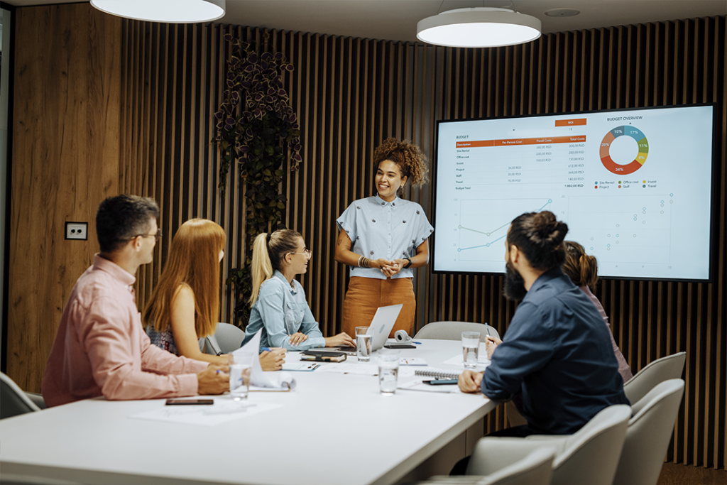 Mulher faz uma apresentação de projeto para um grupo de pessoas sentadas a mesa.