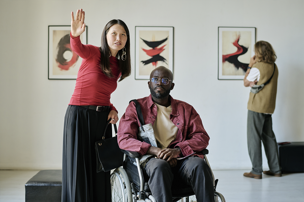Homem cadeirante observando uma exposição de quadros ao lado de uma guia, o que foi possibilitado pela acessibilidade nos eventos